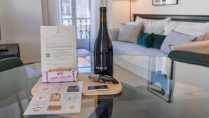 Habitació d'hotel amb taula de vidre, ampolla de vi Ramilo, desenroscador, targeta de benvinguda i fullet informatiu.