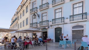 Un café al aire libre con mesas y sillas, un edificio con balcones y un letrero que dice 'O Adriano'.