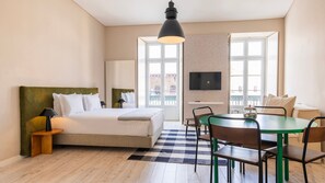 Hotellihuone, jossa on sänky, työpöytä, ruokapöytä tuoleineen, televisio ja suuret ikkunat.