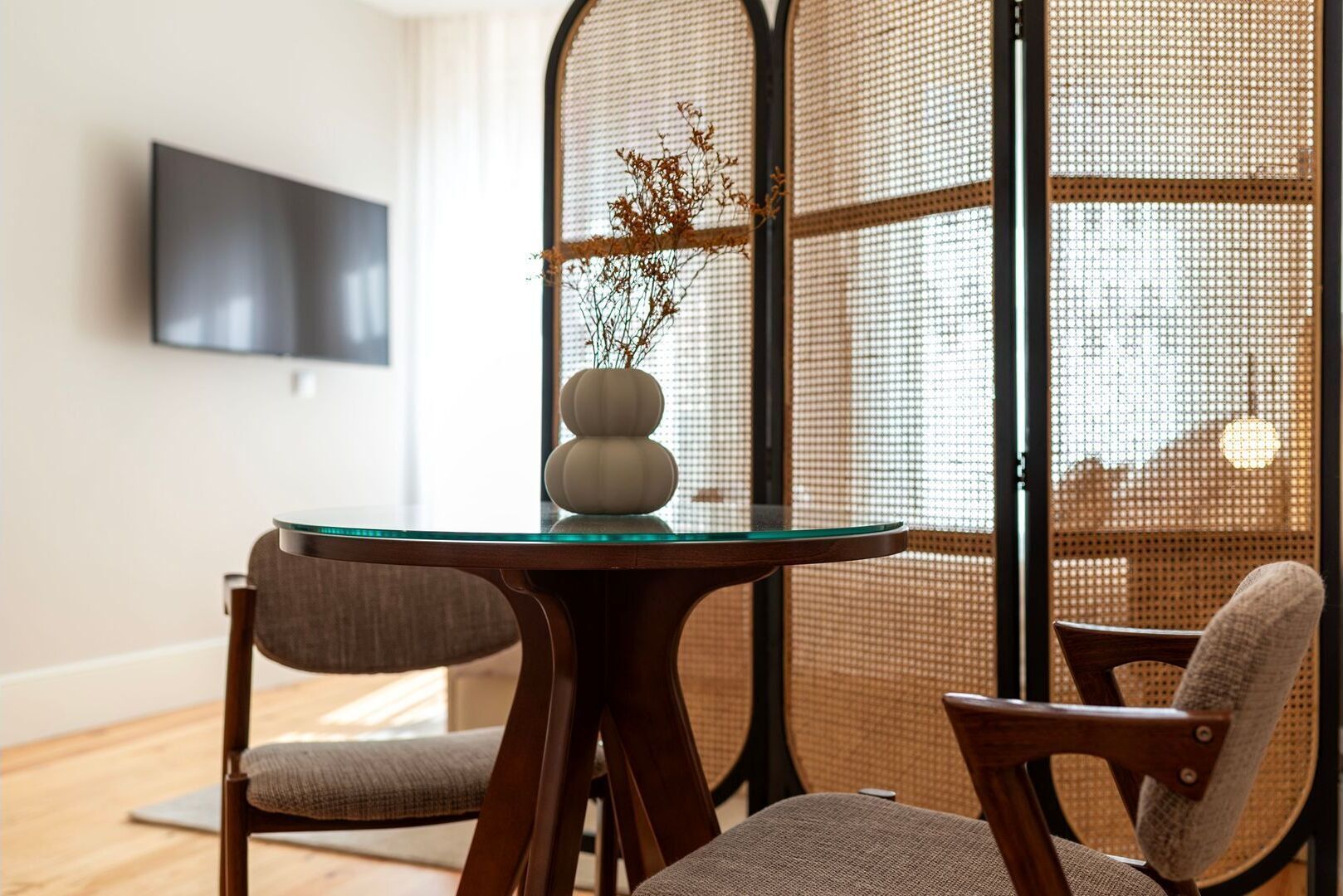 Un comedor moderno con una mesa redonda de cristal, dos sillas tapizadas y un jarrón decorativo con ramas.