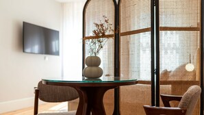 A modern dining area with a round glass table, two upholstered chairs, and a decorative vase with branches. - Ando Living - Flores 200 Townhouse - 102 (Porto)