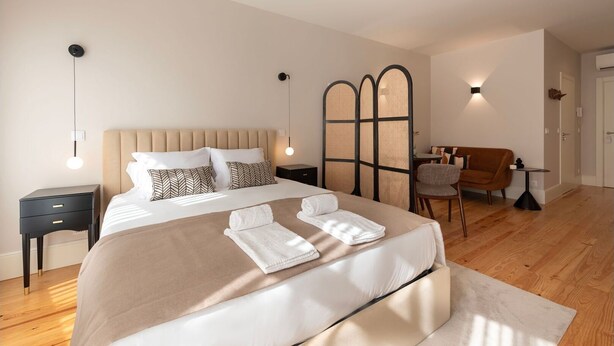 A modern bedroom with a large bed, bedside tables, a seating area with a sofa and armchair, and a partition wall. - Ando Living - Flores 200 Townhouse - 102 (Porto)