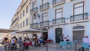 Ein Straßencafé mit Tischen und Stühlen, ein Gebäude mit Balkonen und ein Schild mit der Aufschrift 'O Adriano'.