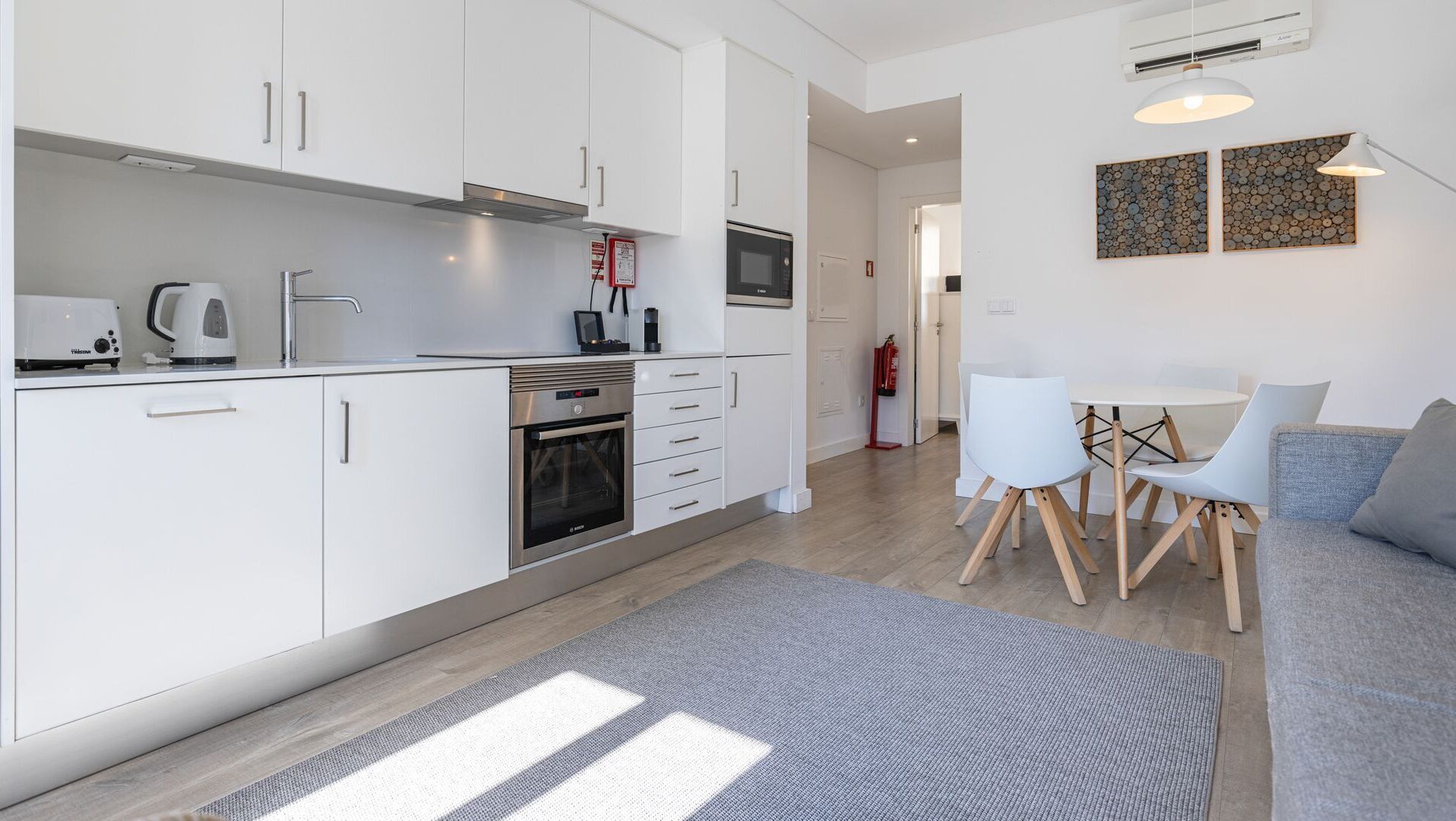 Une cuisine moderne avec des armoires blanches, un four encastré et un four à micro-ondes. On y trouve un coin repas avec une table et des chaises, ainsi qu’un tapis gris sur le sol.