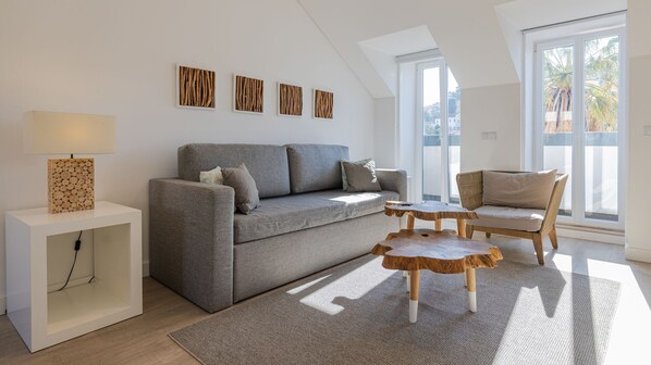 Un soggiorno moderno con un divano grigio, un tavolino in legno, un comodino con una lampada e vista su palme attraverso ampie finestre.
