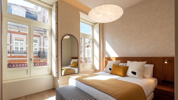 A modern hotel room with a large bed, a wooden headboard, a bedside table, a bench, and a large window with a view of buildings. - Ando Living - Santa Catarina Townhouse - 201 (Porto)