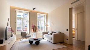 A modern living room with a sofa, a coffee table, a TV, and a chair. There are large windows and a door leading to another room. - Ando Living - Santa Catarina Townhouse - 201 (Porto)