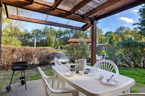 Un patio exterior con una mesa preparada para dos personas, una parrilla y vistas del jardín y la casa circundantes.