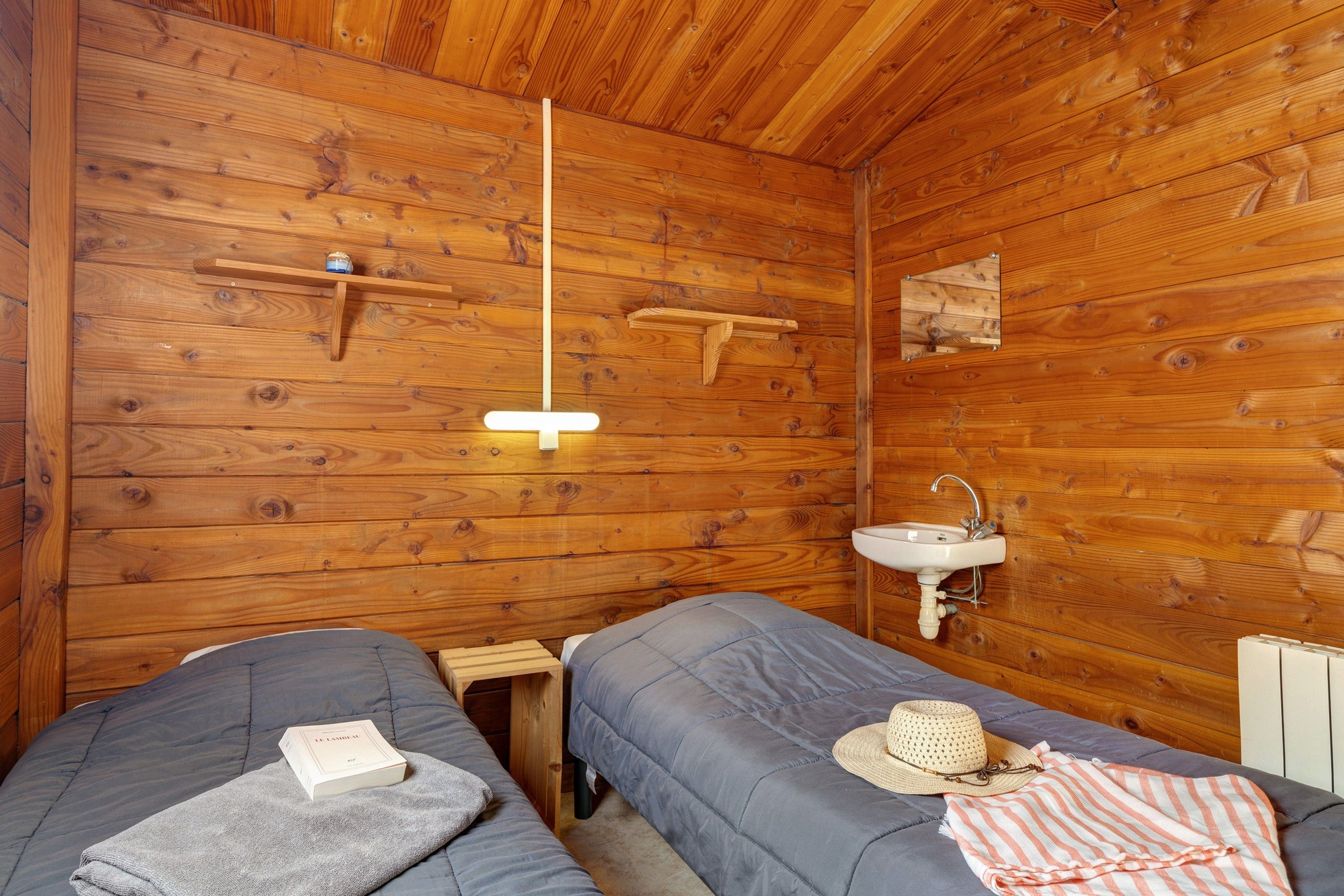 Una habitación tipo cabaña de madera con dos camas, una lámpara colgante, un lavamanos en la pared y un pequeño estante.