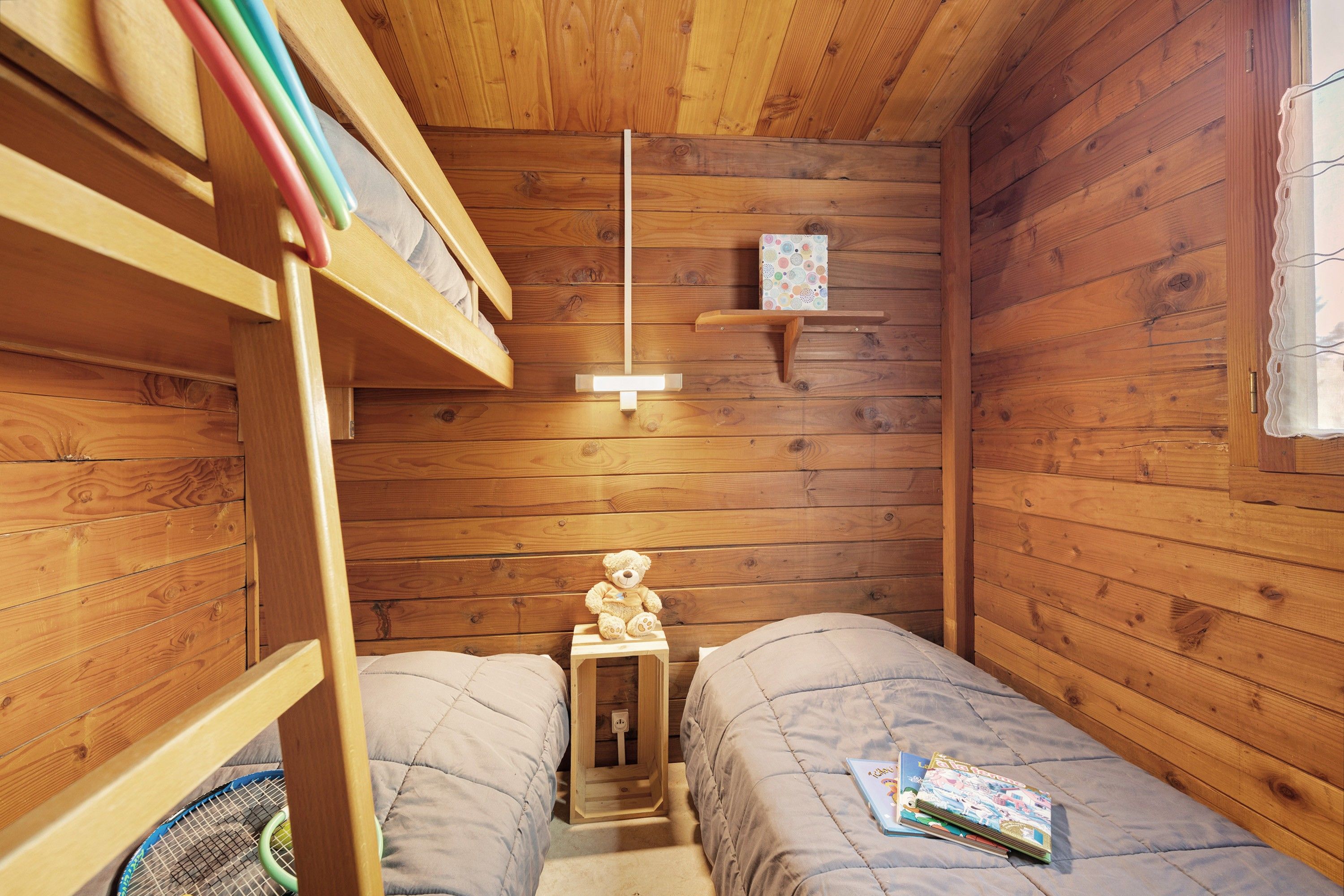 Une petite pièce chaleureuse aux murs en bois, avec un lit superposé. On y trouve une petite table sur laquelle est posé un ours en peluche, ainsi qu’une étagère remplie de livres.