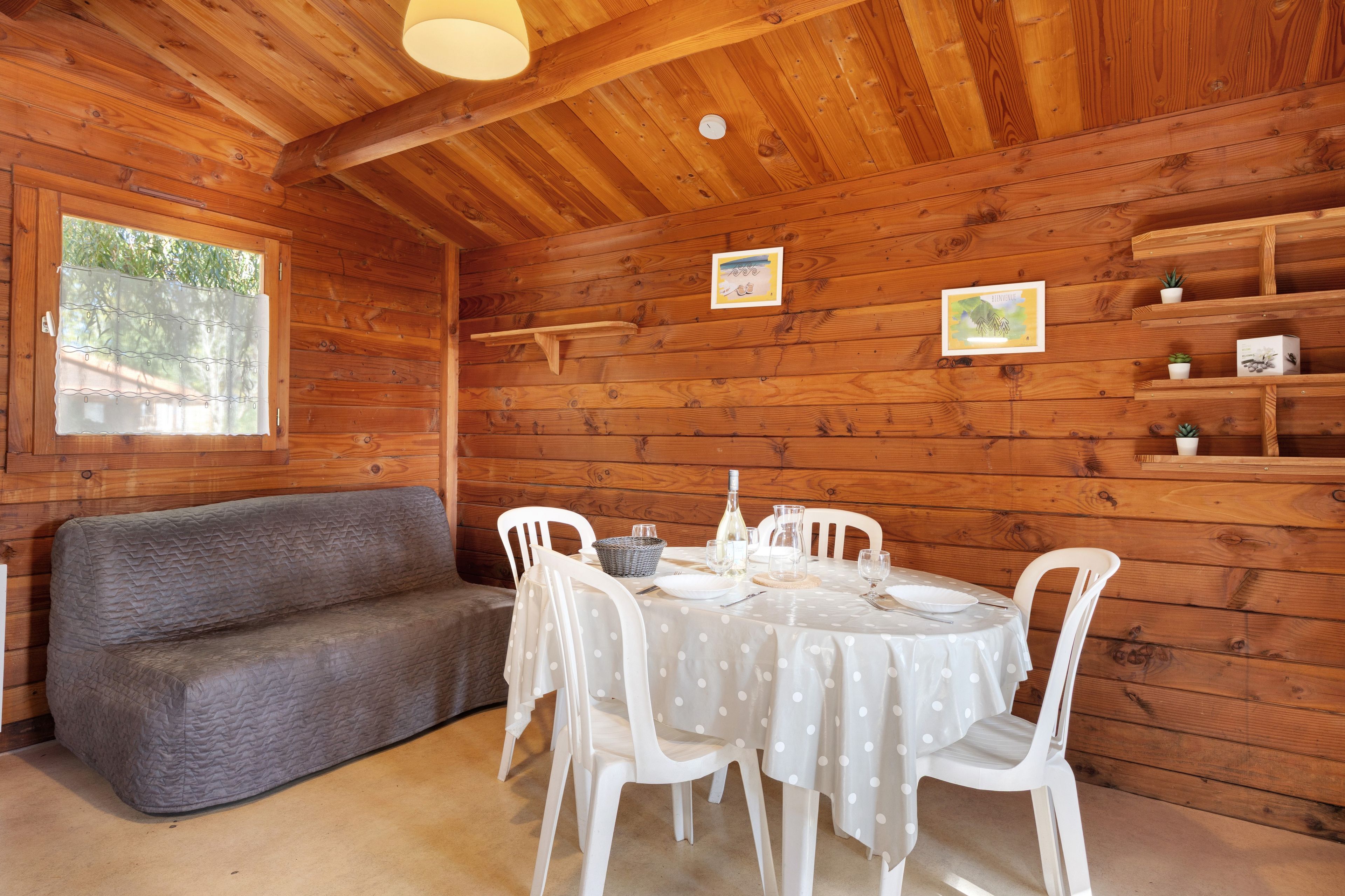L’intérieur d’un chalet en bois, avec une table à manger dressée pour deux personnes, un canapé gris et une fenêtre avec un rideau.