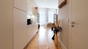 Una cocina moderna con armarios blancos, una cafetera y una lavadora. Hay un comedor con una mesa y sillas, y un jarrón decorativo con plantas secas.
