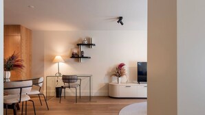 Un salon moderne avec un plancher en bois, une table à manger avec des chaises, une lampe, un téléviseur posé sur un meuble blanc et une petite étagère ornée d’objets décoratifs.