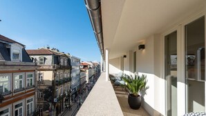 Un balcon étroit orné d’un pot de fleurs, donnant sur une rue bordée de bâtiments historiques et où est garée une voiture.
