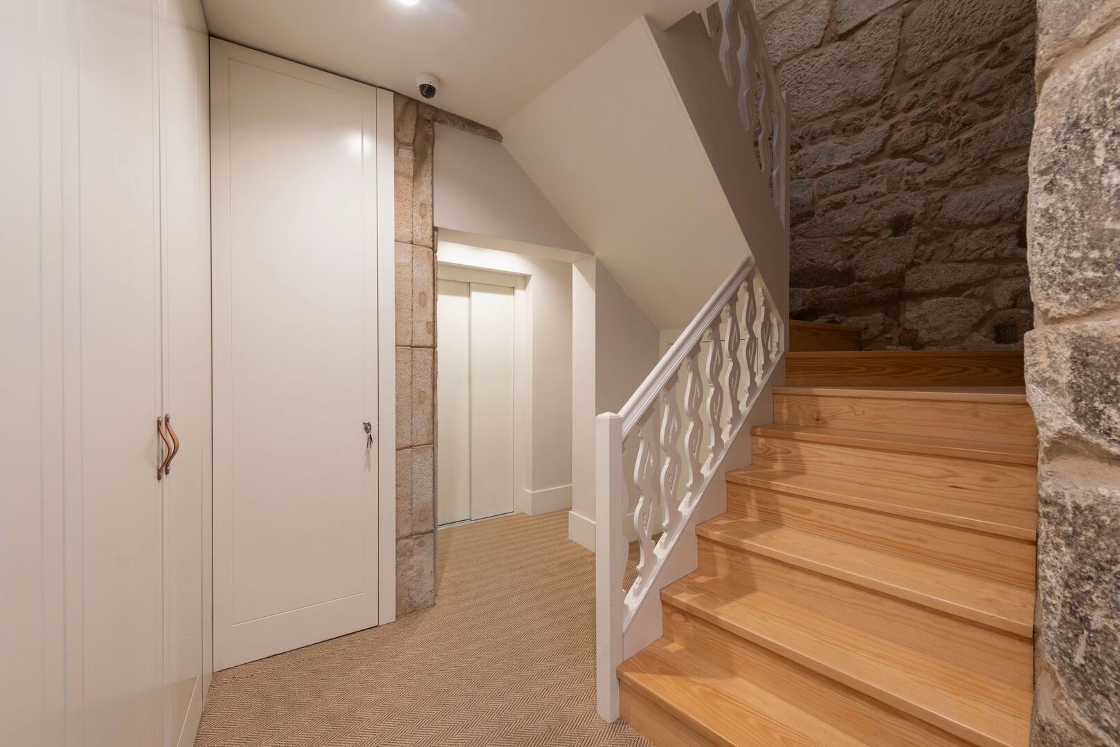Una escalera con escalones de madera y barandillas blancas que conduce a una habitación con pared de piedra y una puerta con cerradura.