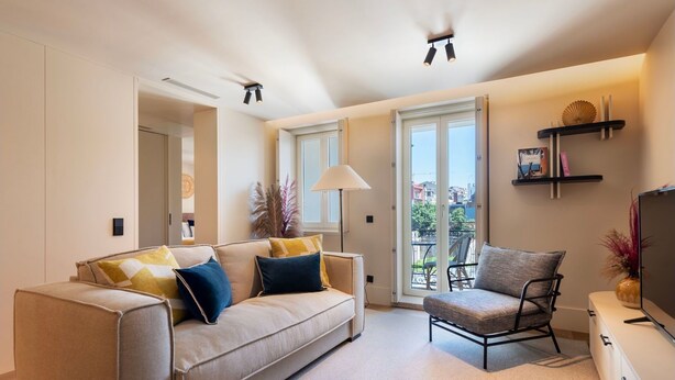 Un salon moderne avec un canapé, un fauteuil et un téléviseur. Il y a un balcon qui offre une vue sur les immeubles.