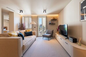 Ein modernes Wohnzimmer mit Sofa, Sessel, Fernseher und Blick auf einen Balkon.