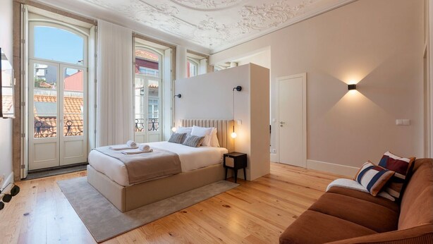 A bedroom with a bed, a nightstand, a sofa, a large window, and a door. - Ando Living - Flores 200 Townhouse - 301 (Porto)