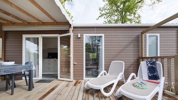 A wooden deck with a table and two lounge chairs, a sliding glass door, and a window. - “Riviera 48” Mobile Home with Shared Pool, Communal Terrace, and Wi-Fi (Saint-Gervais)