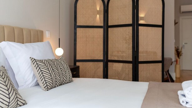 A neatly made bed with white and patterned pillows, a beige headboard, and a bedside table with a lamp. - Ando Living - Flores 200 Townhouse - 302 (Porto)