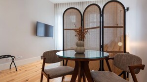 A dining area with a round glass table, four chairs, a vase with dried flowers, and a TV mounted on the wall. - Ando Living - Flores 200 Townhouse - 302 (Porto)