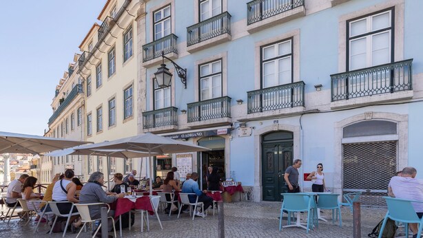 Un café al aire libre con mesas y sillas, un edificio con balcones y un letrero que dice 'O Adriano'.