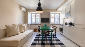 Un espacio habitable compacto con sofá, mesa de comedor, sillas, zona de cocina y televisor montado en la pared.