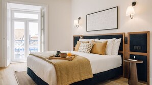A hotel room with a bed, a bedside table, a tray with breakfast items, a wall-mounted lamp, and a framed picture. - Ando Living - Abrantes 50 House - 504 (Lisbon)
