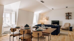 Ein Essbereich mit einem Holztisch, gedeckt für vier Personen, umgeben von Korbstühlen. Im Hintergrund befinden sich ein dunkelblaues Sofa und ein beiger Sessel.