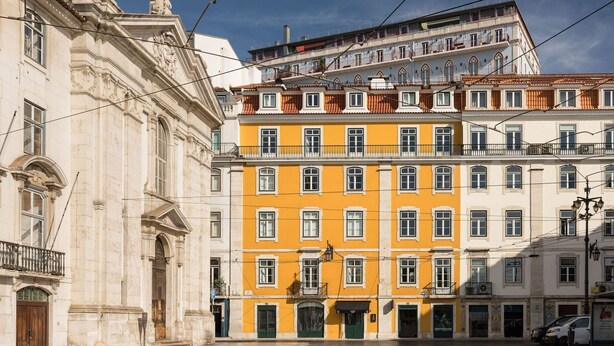 Ein mehrgeschossiges Gebäude mit gelber Fassade, ein Gebäude im klassischen Stil mit weißer Fassade und eine Straße mit geparkten Autos.