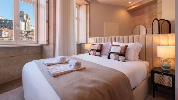 A hotel room with a large bed, a nightstand with a lamp, and a view of a historic building through the window. - Ando Living - Flores 200 Townhouse - 303 (Porto)