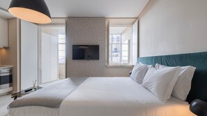 A modern bedroom with a large bed, a flat-screen TV mounted on the wall, and a view of the city through a window. - Carmo 43 | Apartment 400 (Lisboa)
