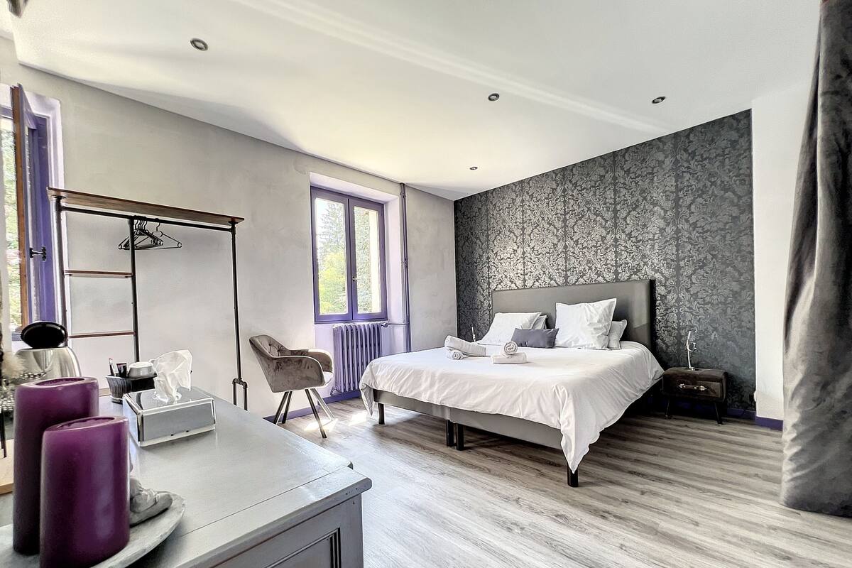 A modern bedroom with a large bed, a chair, a dresser, and a window with a view of trees.