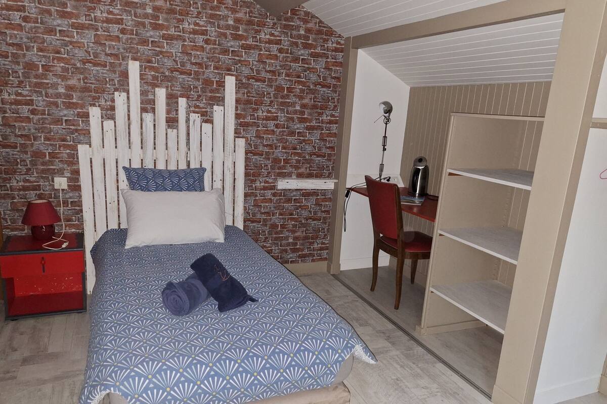 A bedroom with a brick wall, a bed with a patterned bedspread, a red nightstand, a red chair, and a desk with a kettle.
