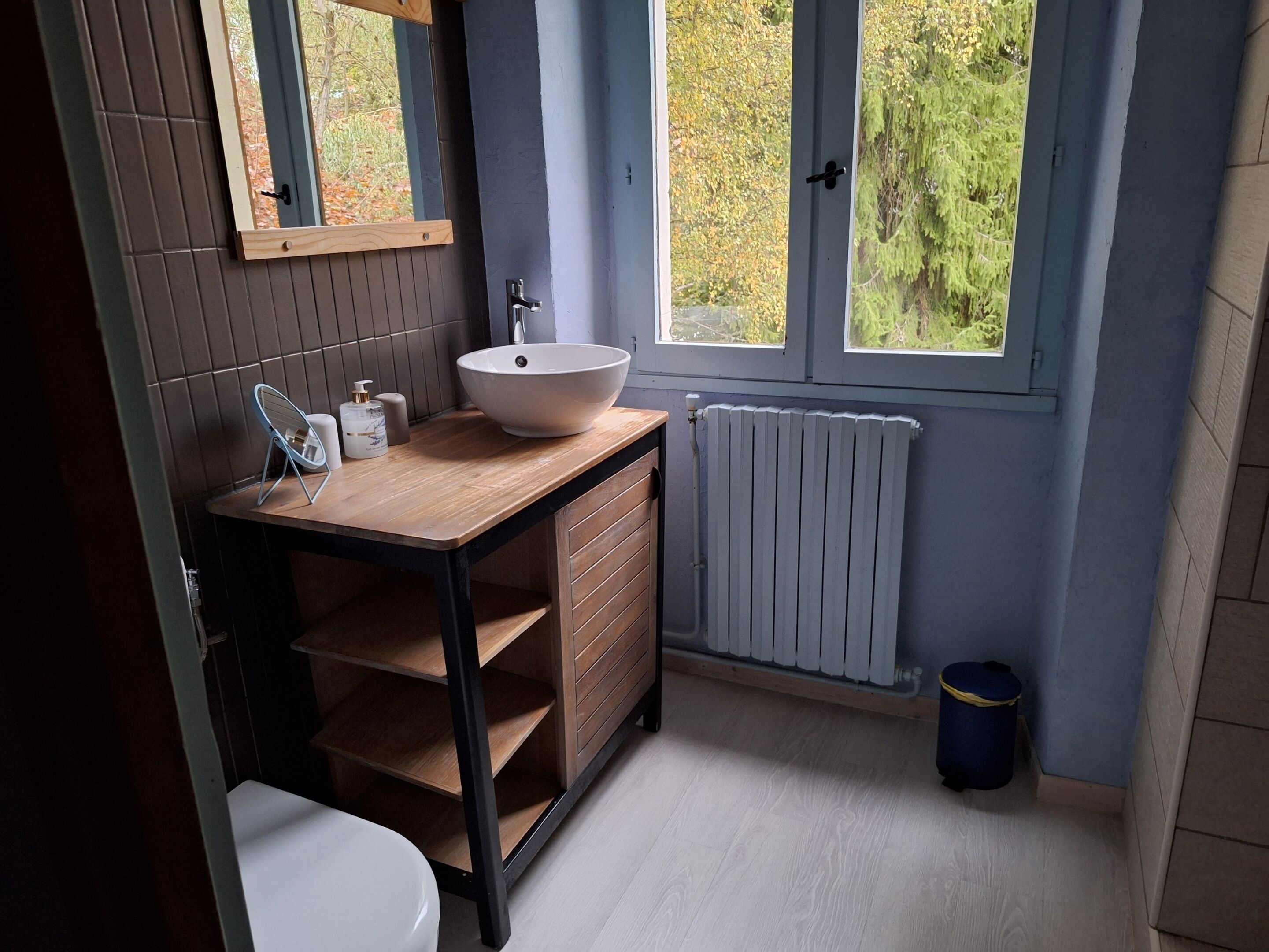 Un baño moderno con tocador de madera, lavamanos blanco y ventana con vistas a árboles.