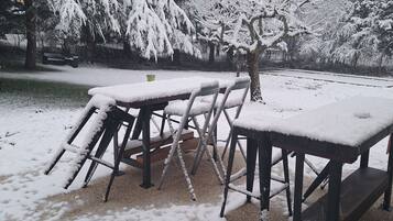 Un área de estar al aire libre cubierta de nieve, con mesas y sillas, rodeada de árboles cargados de nieve.