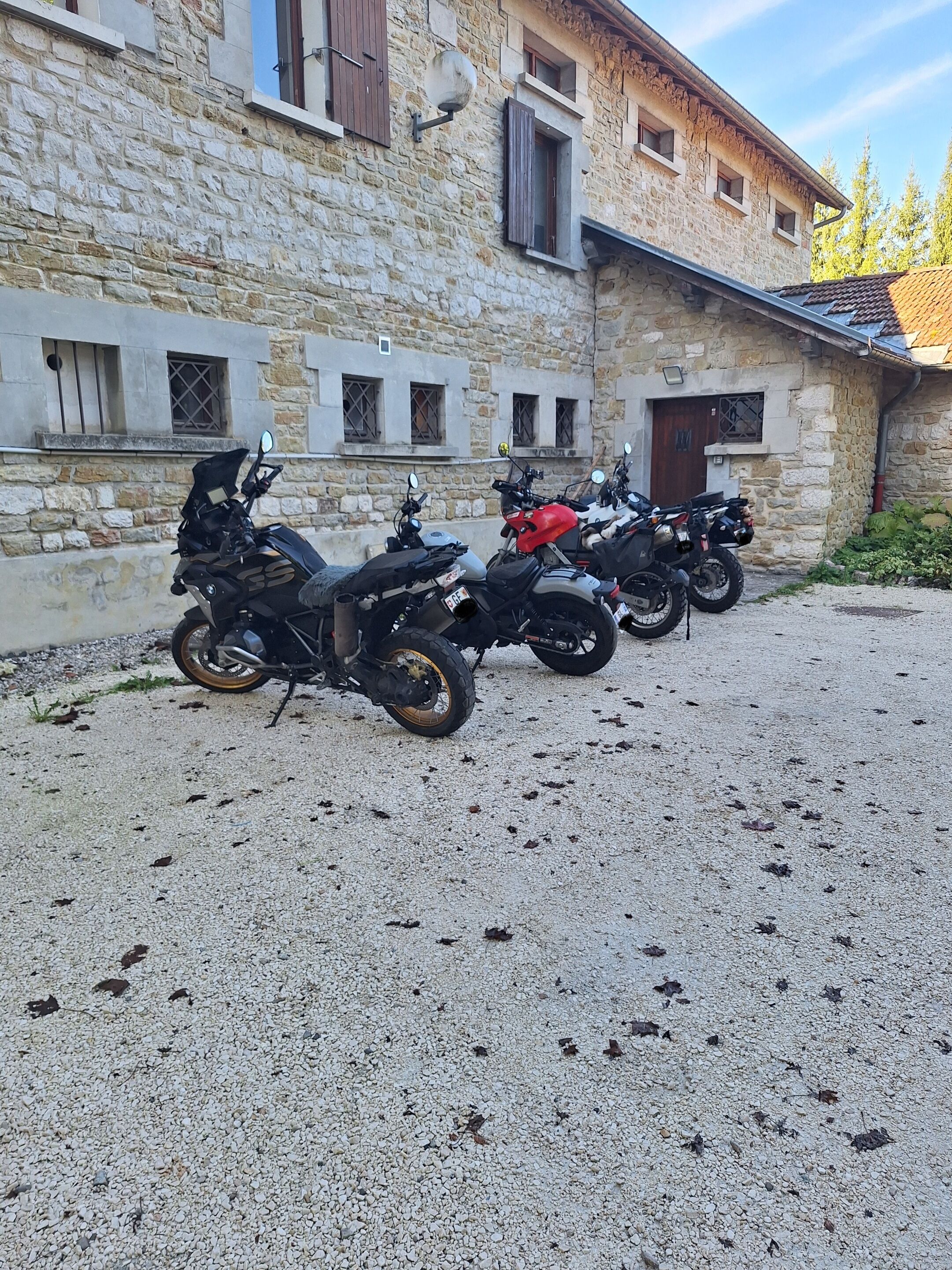 A row of motorcycles parked in front of a stone building with multiple windows and a gravel ground.