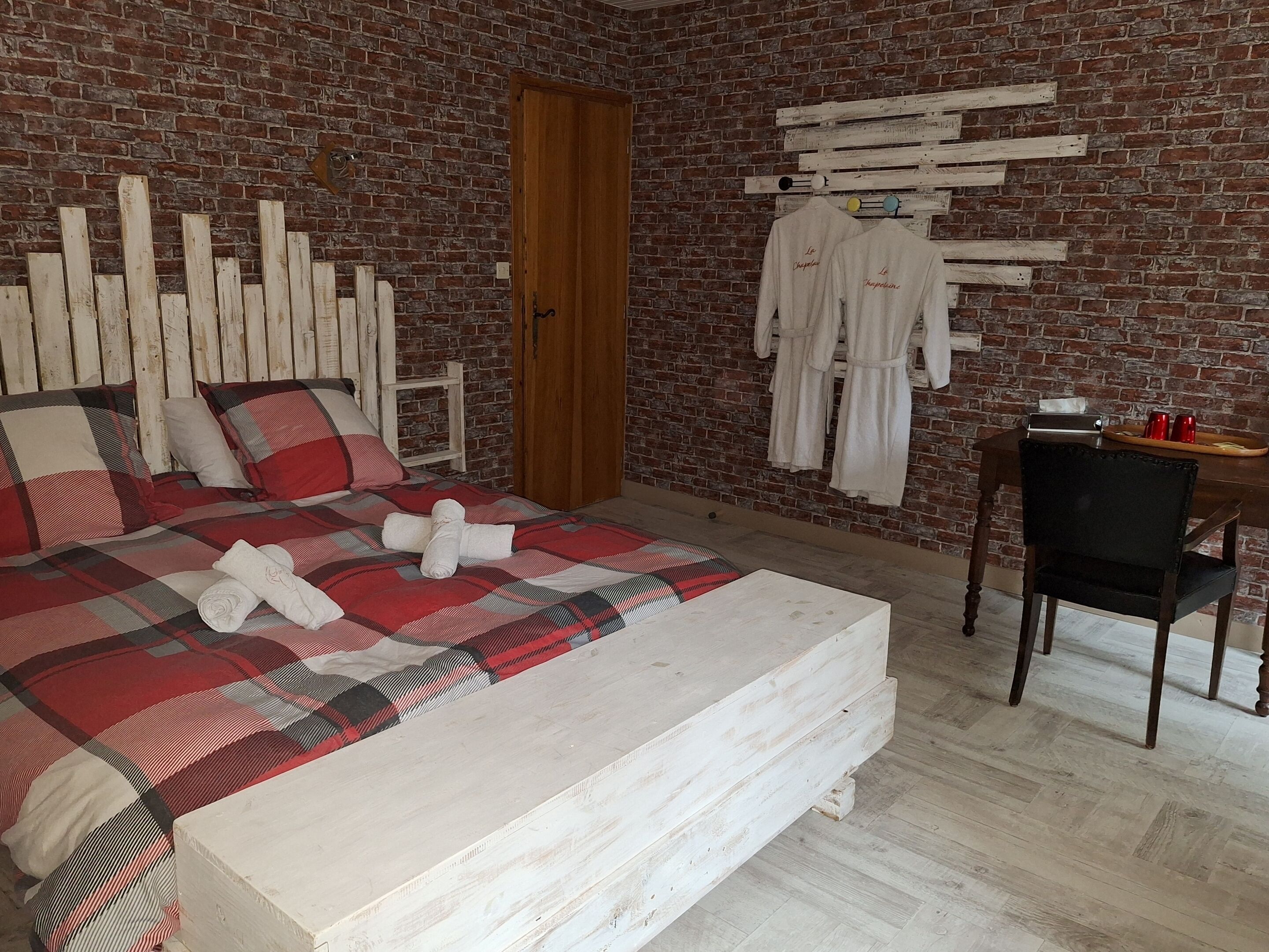Un dormitorio con pared de ladrillo, estructura de cama de madera, mesita de noche de madera, una silla y un escritorio de madera con una bandeja de tazas.