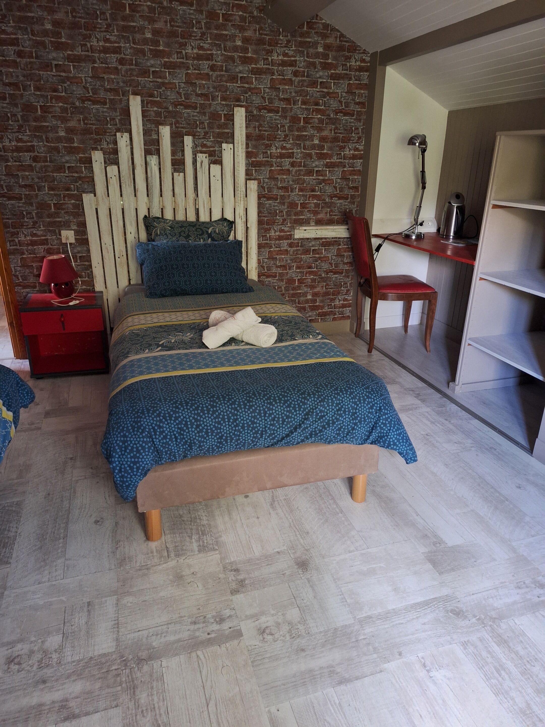 Un dormitorio con pared de ladrillo, cabecera de madera, cama con colcha azul estampada, mesita de noche roja y un escritorio con silla.