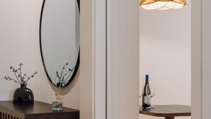 Une pièce avec un grand miroir ovale, un petit vase contenant des branches, une bouteille de vin et une table avec des verres.
