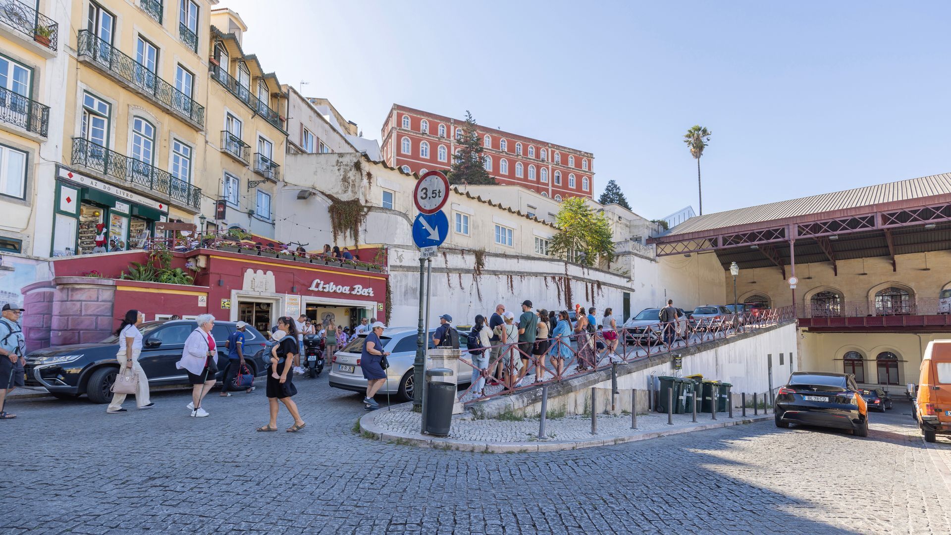 Uma rua de paralelepípedos com uma multidão de pessoas, um bar chamado 'Lisboa Bar', e um prédio vermelho com uma placa '3,5f'.