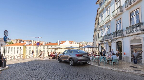 Una calle adoquinada con un coche aparcado, mesas al aire libre en cafés y edificios históricos con balcones.
