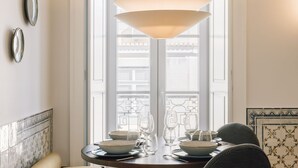 Et spiseområde med et rundt bord dækket til to personer, en hængende lampe og udsigt gennem et vindue med dekorativt jerngitter.