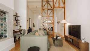 Un salon moderne avec un canapé vert, une table basse et un téléviseur posé sur un meuble en bois. Une structure en poutres apparentes se trouve au centre de la pièce.