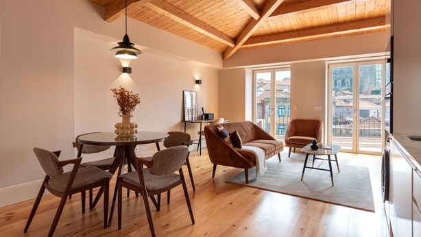 Um quarto espaçoso com teto de madeira, área de jantar com mesa redonda e cadeiras, sofá e vista para edifícios através de grandes janelas.