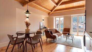 Una habitación amplia con techo de madera, un comedor con mesa y sillas redondas, un sofá y vistas a edificios a través de ventanas grandes.