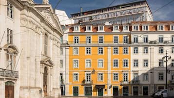 Un immeuble à plusieurs étages avec une façade jaune, un bâtiment blanc à l’architecture classique, et une rue bordée de voitures garées.