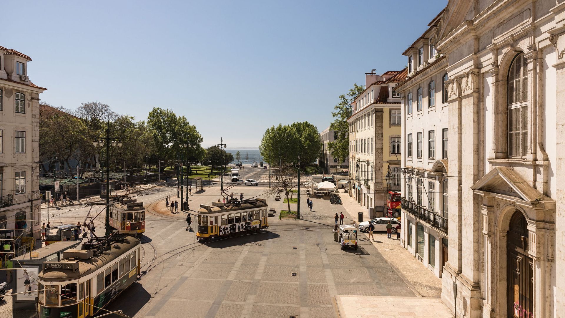 A historic square with tram lines, buildings, and a clear view of the sea.