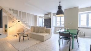 A modern living room with a staircase, a sofa, a dining table with chairs, and a large window. - Carmo 43 | Apartment 403 (Lisboa)