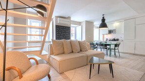 A modern living room with a sofa, a coffee table, and a chair. There is a staircase with a railing, a dining area with a table and chairs, and a kitchen area with a microwave and cabinets. - Carmo 43 | Apartment 403 (Lisboa)